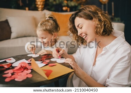 Mother and little daughter making Valentine's day cards using color paper, scissors and pencil, sitting by the table in cozy room  Royalty-Free Stock Photo #2110040450