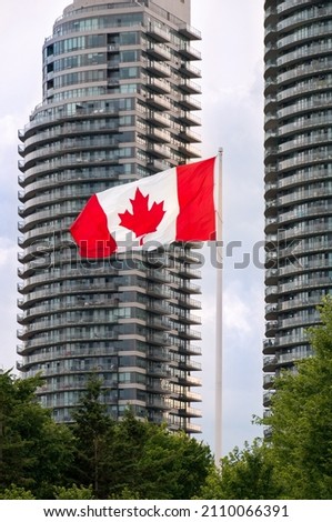 Red and white Canadian flag waving on the mast above the park trees in front of modern high-rise buildings. Red maple leaf is a state symbol of Canada depicted on Canadian flag.