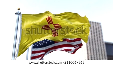 the flag of the US state of New Mexico waving in the wind with the American flag blurred in the background. New Mexico state flag It is one of four U.S. state flags not to contain the color blue Royalty-Free Stock Photo #2110067363