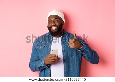 Technology and online shopping concept. Happy african-american guy showing thumb-up in approval while using mobile phone, standing satisfied over pink background