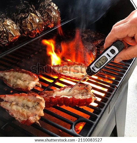 A person checks the temperature while cooking lobster tail and potatoes on a barbecue.