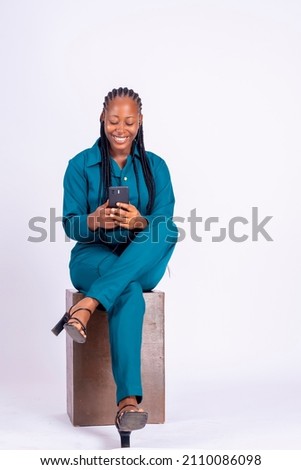 Full length shot of a pretty young woman sitting in an easy chair, looking at her phone screen, using a new trendy application against a white studio wall.  Royalty-Free Stock Photo #2110086098