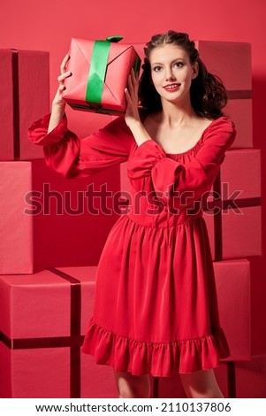 Charming girl with evening makeup and hairstyle in a nice red dress holds joyfully a gift box thinking what is inside of it. Bunch of red gift boxes background. Christmas and New Year presents. 
