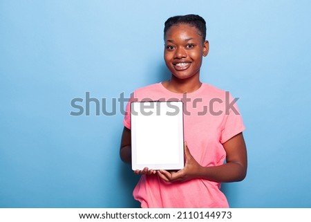 Portrait of african american young woman holding tablet computer with white screen standing in studio with blue background. Teenager recommending advertisement ipad application. Isolated touchpad