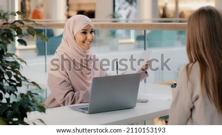 Young arab woman consultant sales agent bank worker sitting at office desk advises unrecognizable girl client offers to sign contract sale agreement helps to arrange loan proposes legal consultation Royalty-Free Stock Photo #2110153193