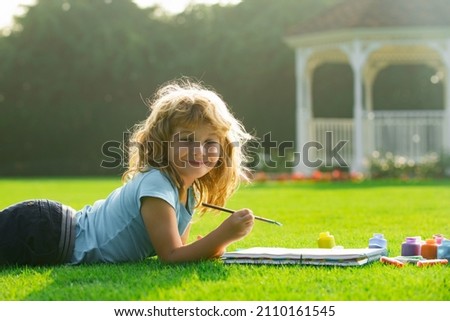 Child boy draws in park laying in grass having fun on nature background. Children artist paints creativity vacation, kids crafts.