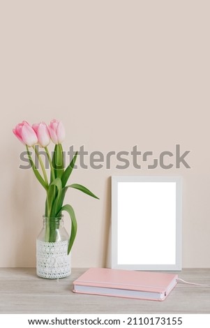 Home interior with decorative elements. Layout with a white frame and pink tulips in a vase and a pink notebook on the table on a light beige background