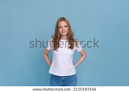 Women's beauty. Elegant attractive model in casual white t-shirt and jeans standing with folded hands behind her back and looking at camera with flirtatious smile, isolated on blue studio background Royalty-Free Stock Photo #2110183166