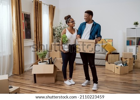 Couple in love moves into new apartment, they carry cardboard boxes with packed belongings into living room, woman sets up decorations, holds pot with plant in hand they decorate first home together Royalty-Free Stock Photo #2110223459