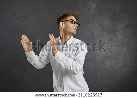 Cheerful young man in modern thug life glasses dancing, enjoying music and having fun. Happy positive guy in pixel sunglasses doing funny dance moves isolated on grey background