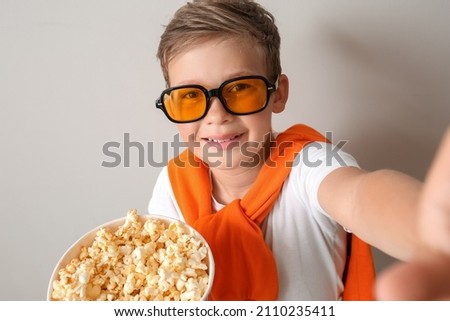 Little boy in eyeglasses with bucket of tasty popcorn taking selfie on light background