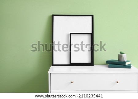 Blank frames and books on chest of drawers near green wall