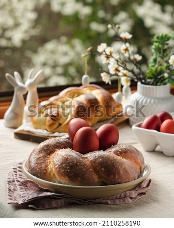 Easter traditional bread, austrian ostern zopf, greek tsoureki and red eggs on a table with linen tablecloth with spring window view, still life Royalty-Free Stock Photo #2110258190