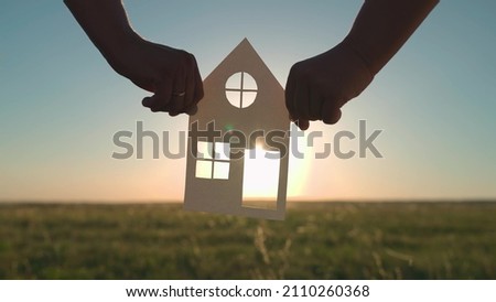 The family's hands are holding a paper house at sunset, the sun is shining through the window. The symbol of the house, happiness. The concept of building a house for the family. Dream to buy a house