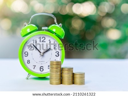 Green alarm clock and stacks of golden coins on blur green plant background. Concept of time is money concept