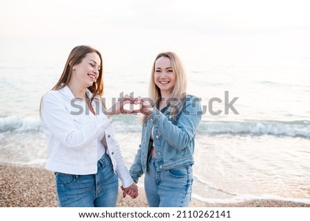 Happy two women show heart shape with hands posing over sea background outdoor. Friends spending time together. Summer holiday season. Valentines Day 