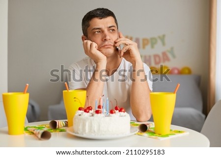 Portrait of sad pensive young man wearing white casual style T-shirt sitting at table with cake, talking smart phone with her friend, celebrating birthday alone, feeling sorrow.