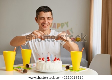 Indoor shot of smiling positive brunette man wearing white casual style T-shirt sitting at table with cake, taking picture of his festive food, expressing happiness.