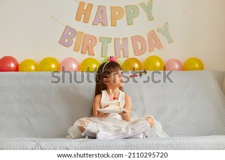 Image of happy little girl wearing white dress sitting on sofa, blowing party horn and holding cake with burning candle, looking away, child celebrating birthday.