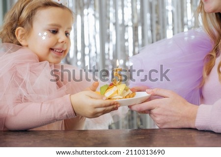 Preschool fair-haired girl having festival pink dress and stars decoration on her face holding cake with candle on her 5th birthday anniversary in cafe with decorated background of silvery spangle