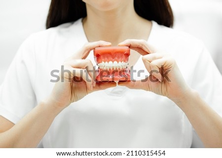 Young unrecognizable woman doctor dentist orthodontist with black hair in white medical clothes holding tooth model with wire braces attached in two hands. Dental care, medical, oral hygiene concept. Royalty-Free Stock Photo #2110315454