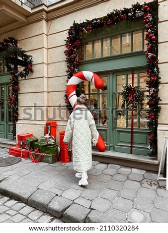 blonde girl dressed in white quilted down jacket, milky massive shoes and scarf, fairytale house with Christmas decor on background, doors with Christmas tree, red toys, gift boxes and lollipop