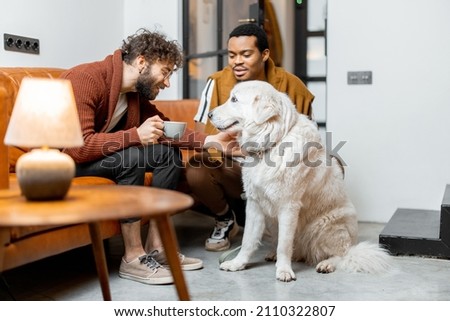 Men care their huge white dog at cozy home. Concept of homosexual relations and lifestyle at home. Idea of multinational gay families Royalty-Free Stock Photo #2110322807