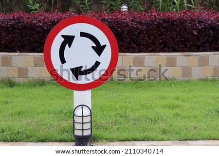 White and red roundabout sign on the road with plants in park in the village background. Sign and transportation concept.