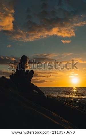 Silhouette of a young woman sitting on huge stones on the seashore and looking at a beautiful, magical, bright sunset with the sun going over the sea. Vertical photo