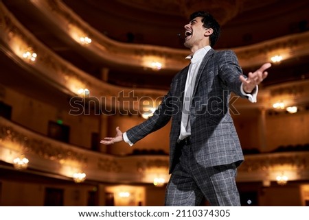 professional opera singer in elegant classic outfit, singing hit with open mouth in microphone gesture with hand standing on stage in theater. Handsome caucasian guy during performance Royalty-Free Stock Photo #2110374305