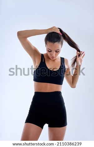 portrait of slim fitness woman making ponytail preparing for sport workout, wearing bblack tracksuit short top and shorts. brunette lady is looking down confidently. sport and fitness concept Royalty-Free Stock Photo #2110386239