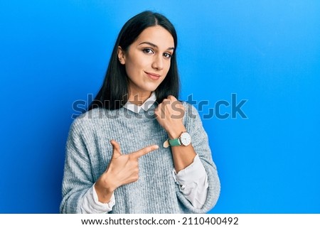 Young hispanic woman wearing casual clothes in hurry pointing to watch time, impatience, looking at the camera with relaxed expression  Royalty-Free Stock Photo #2110400492