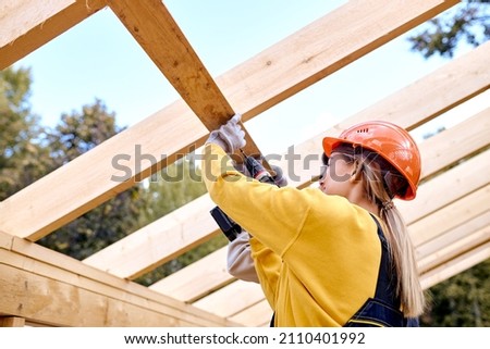 Two Positive Contractors Working With Drill Driver. Carpenter Working on Wooden House Skeleton Frame Roof Section. Construction Industry Theme. Woman In Hardhat Is Laughing During Work Royalty-Free Stock Photo #2110401992