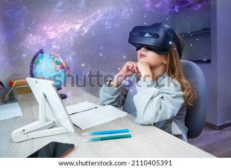 Schoolgirl using VR headset simulators for studying. Child is wearing virtual reality glasses sitting at desk with tablet and watching on sky of stars. Future learning concept Royalty-Free Stock Photo #2110405391