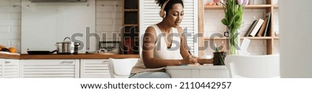 Black woman in headphones working with laptop while sitting at table in home kitchen