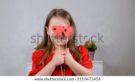 funny Pretty little girl in a red dress with two hearts on a stick closes her eyes with them. Valentines day concept.