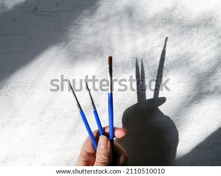 In a woman's hand, blue brushes for drawing cast a shadow on the canvas with the drawing by numbers.