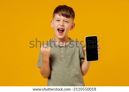 Little happy fun boy 6-7 years old in green t-shirt hold use mobile cell phone with blank screen workspace area do winner gesture isolated on plain yellow background Mother's Day love family concept