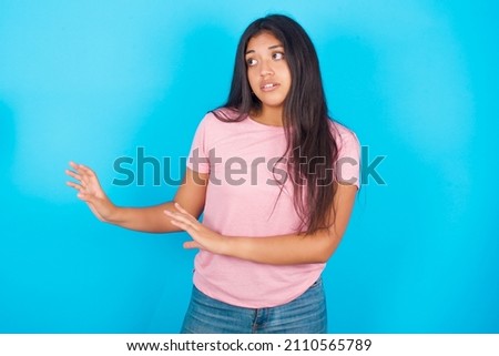 Displeased Young beautiful woman wearing pink T-shirt against blue background keeps hands towards empty space and asks not come closer sees something unpleasant