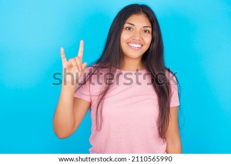 Young beautiful woman wearing pink T-shirt against blue background doing a rock gesture and smiling to the camera. Ready to go to her favorite band concert.