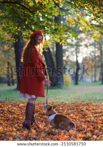 A girl with long red hair, in a red coat and beret is training her corgi dog in the autumn park. Golden autumn, walking with a dog, friendship, dancing with a dog. beautiful autumn picture