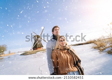 Loving couple having fun on ice in typical dutch landscape with windmill. Woman and man ice skating outdoors in sunny snowy day. Romantic Active date on frozen canal in winter Christmas Eve.