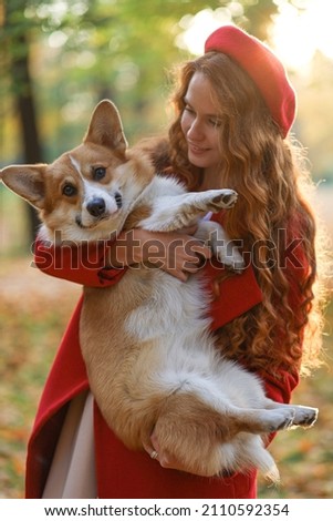 A girl with long red hair, in a red coat and beret gently hugs her corgi dog in the autumn park. Golden autumn, walking with a dog, friendship, dancing with a dog. beautiful autumn picture