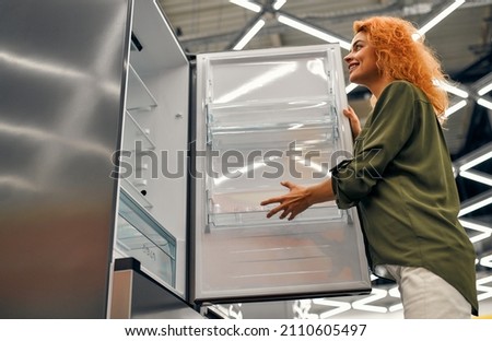 Beautiful red-haired woman choosing a refrigerator in a home appliances and electronics store. Buying a new refrigerator.