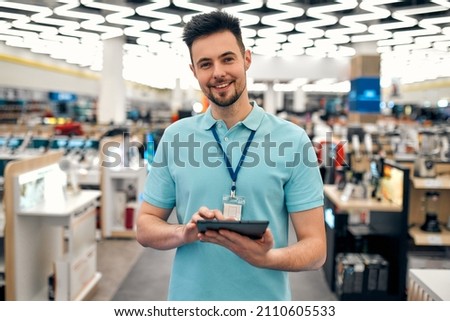 A young successful sales assistant in uniform with a tablet in his hands against the background of the interior of the hall in a home appliances and electronics store. Royalty-Free Stock Photo #2110605533