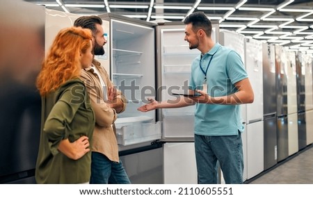 Beautiful red-haired woman and bearded man buying a refrigerator in a home appliances and electronics store. Male sales assistant helping a couple choose new appliances for the home.