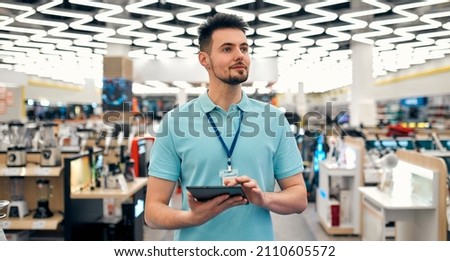 A young successful sales assistant in uniform with a tablet in his hands against the background of the interior of the hall in a home appliances and electronics store. Royalty-Free Stock Photo #2110605572
