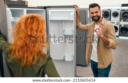 A young couple is choosing a new refrigerator in a home appliances and electronics store. A man with a beard shows an ok gesture while holding on to the refrigerator door.