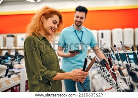 A beautiful red-haired girl is talking to a male salesperson while getting help buying a new hair straightener at a hardware and electronics store. Royalty-Free Stock Photo #2110605629