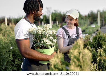 Excited Caucasian and black farmers standing in greenhouse talking, taking notes. Bearded black man holding flower plant in pot,redhead woman with paper, writing, talking. Commercial gardening Royalty-Free Stock Photo #2110620041